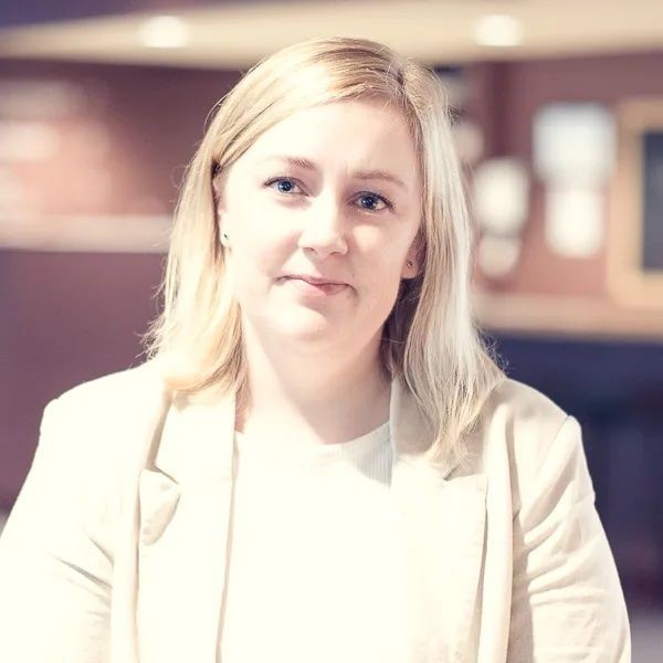 Portrait of a person with blonde hair and a light beige blazer in a softly lit interior background.